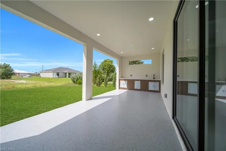 Expansive covered patio with a speckled concrete finish, recessed lighting, and a built-in outdoor kitchen featuring wood-finish cabinetry and a dark faucet