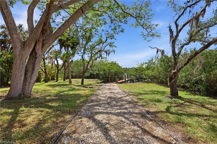 The property features a gravel pathway leading to a wooden dock, surrounded by mature trees and lush green vegetation under a clear blue sky