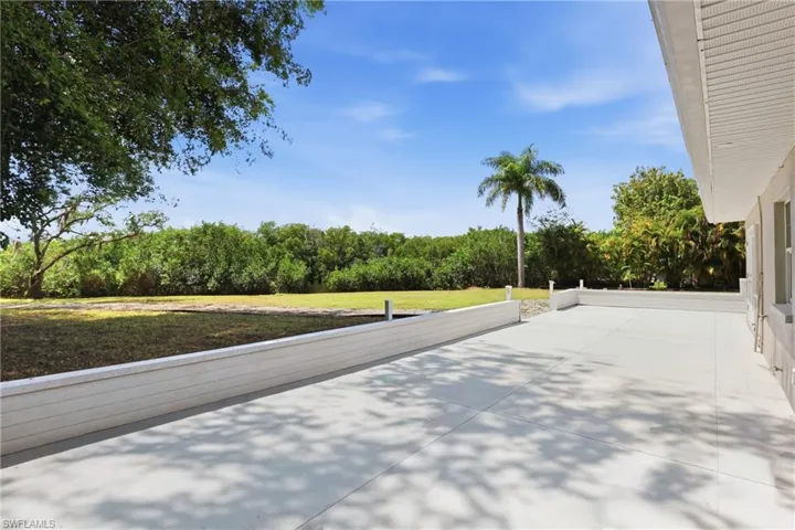 Expansive outdoor patio area featuring a concrete surface and a low white retaining wall, offering views of a spacious grassy area with mature trees and tropical foliage