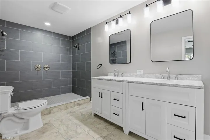 This bathroom features a spacious shower with gray tile walls, a white dual vanity with marble countertops, and two rectangular mirrors
