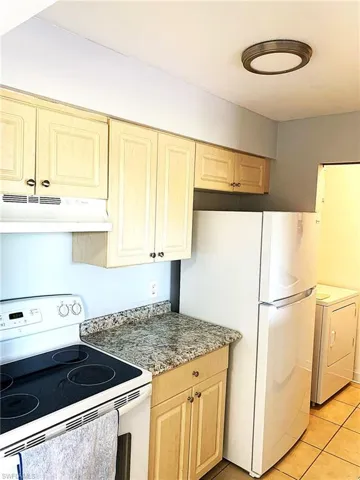 Kitchen with white appliances, under cabinet range hood, light tile patterned flooring, light stone countertops, and light brown cabinetry