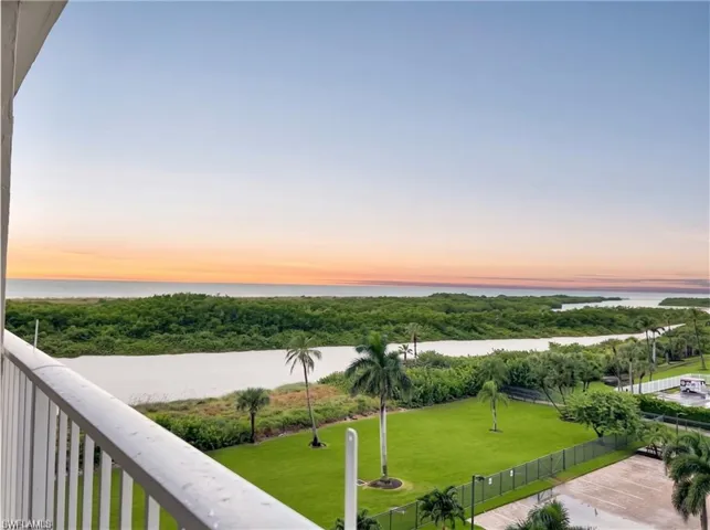 Balcony featuring white metal railings, overlooking a manicured lawn, palm trees, a water feature, and a horizon with water