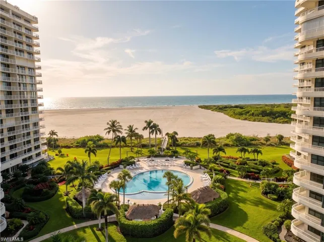 Beachfront pool area featuring mature palm trees, manicured landscaping, paved walkways, and thatched-roof structures