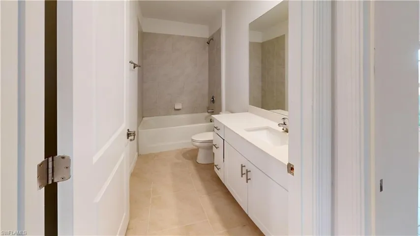 Bathroom featuring a white vanity with integrated sink, a rectangular mirror, a bathtub with tiled surround, a toilet, and light-toned floor tiling