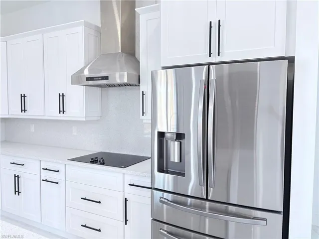 Kitchen with wall chimney range hood, white cabinetry, light stone counters, black electric cooktop, and stainless steel fridge with ice dispenser