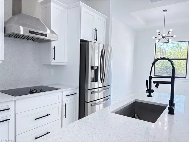 Kitchen with stainless steel refrigerator with ice dispenser, wall chimney exhaust hood, sink, light stone countertops, and white cabinets