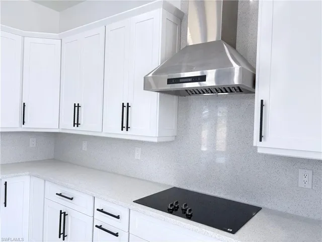 Kitchen featuring black electric cooktop, wall chimney range hood, backsplash, and white cabinets