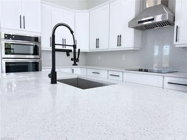 Kitchen with black electric stovetop, wall chimney range hood, white cabinets, and light stone counters