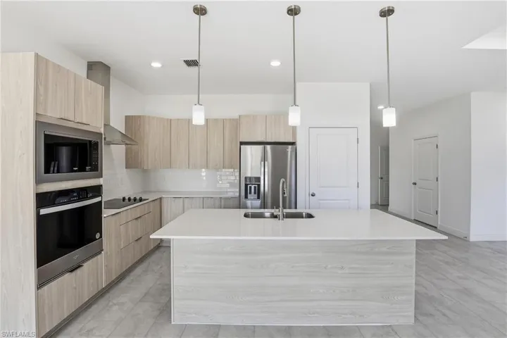 Kitchen with modern cabinets, light brown cabinetry, pendant lighting, stainless steel appliances, and wall chimney exhaust hood