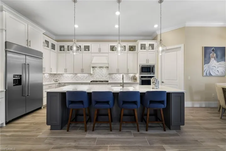 Kitchen with a center island with sink, custom exhaust hood, built in appliances, backsplash, and decorative light fixtures