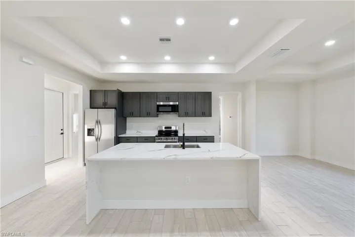 Kitchen featuring light stone counters, a center island with sink, a raised ceiling, and recessed lighting