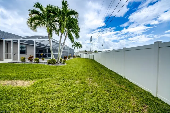 Screened-in pool enclosure, mature palm trees, manicured lawn, established landscaping, and a white privacy fence