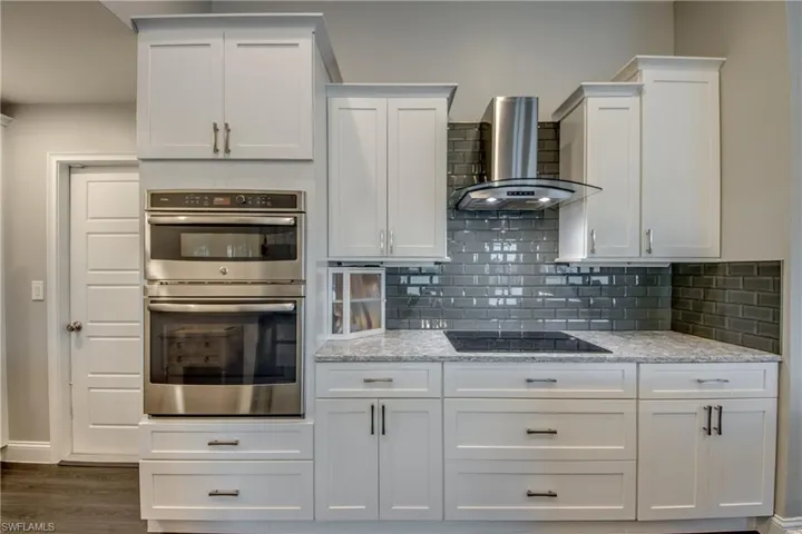 White shaker-style cabinetry, double wall ovens, built-in cooktop, stainless steel range hood, and speckled countertops