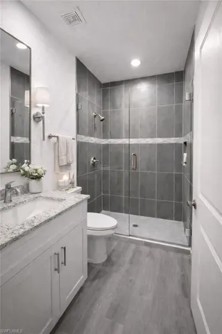 Bathroom featuring a frameless glass shower enclosure with dark gray subway tile, a white vanity with a granite countertop, and wood-finish flooring
