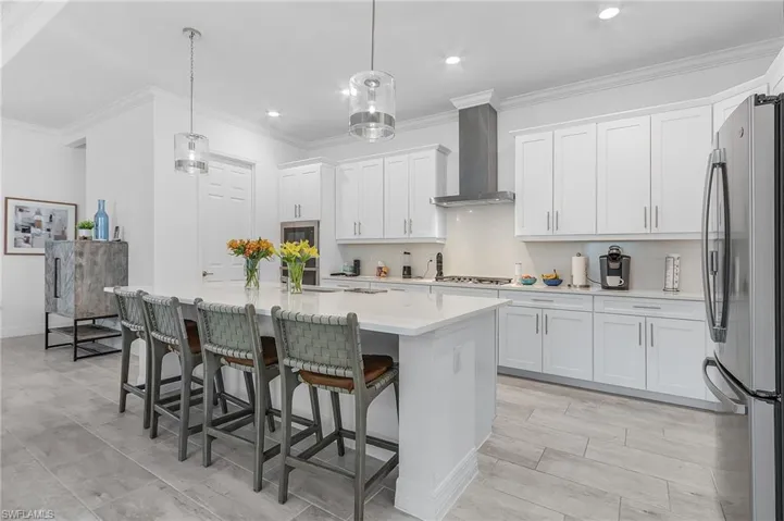 Kitchen with white cabinets, stainless steel fridge, a kitchen island with sink, wall chimney exhaust hood, and decorative light fixtures