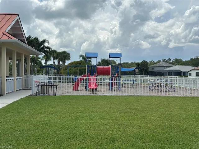 Fenced playground with colorful equipment, sand ground cover, adjacent grassy area, and a covered pavilion with a red metal roof