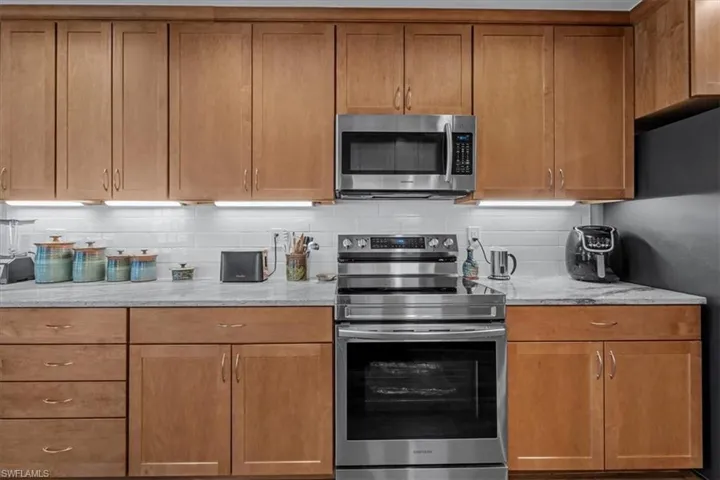 Kitchen featuring light wood cabinetry with brushed nickel hardware, light-toned countertops, a white subway tile backsplash, under-cabinet lighting, and stainless steel appliances including an oven range and microwave