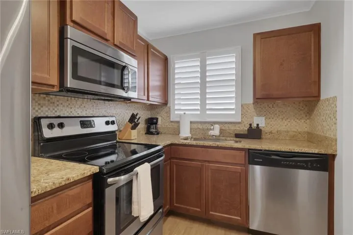 Kitchen featuring wood cabinetry, stainless steel appliances, granite-finish countertops, mosaic tile backsplash, and a window with white plantation shutters