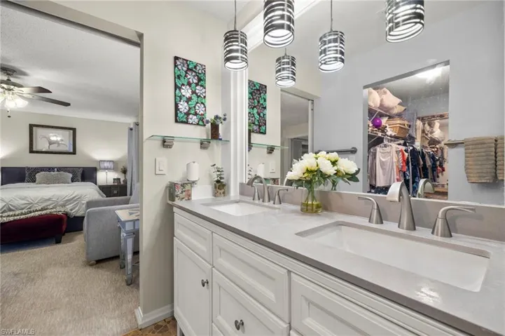 Full bath with double vanity, leads to primary bedroom, a walk in closet, light carpet, and a ceiling fan