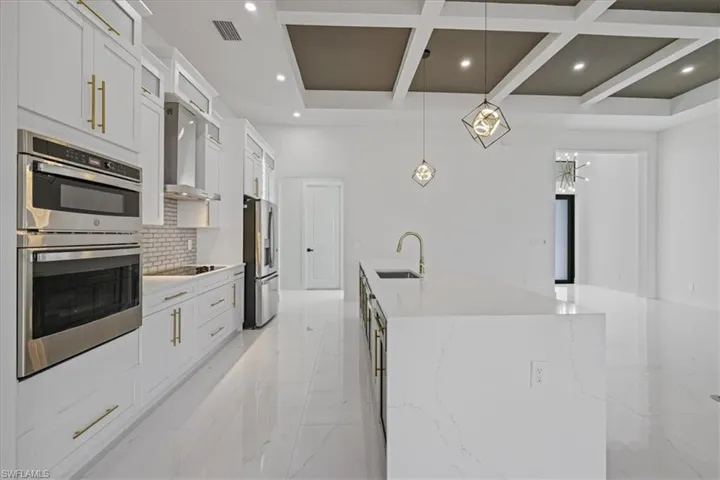 Kitchen with stainless steel appliances, hanging light fixtures, light stone countertops, coffered ceiling, and white cabinetry