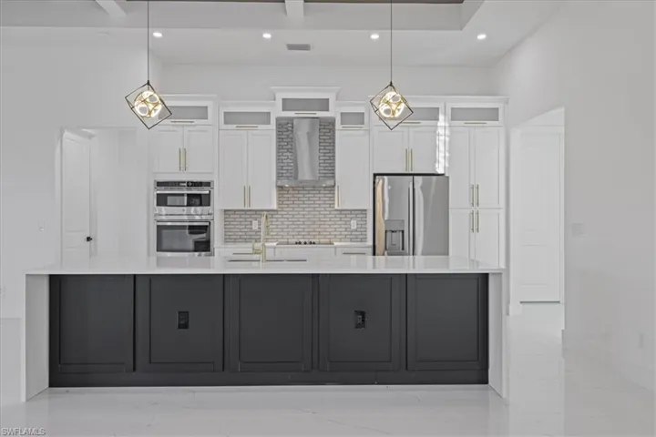 Dual tone kitchen featuring light stone countertops, backsplash, decorative light fixtures, stainless steel appliances, and a spacious island