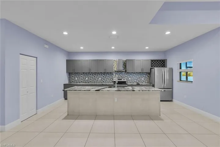 Kitchen with light stone countertops, wall chimney exhaust hood, stainless steel appliances, an island with sink, and gray cabinets