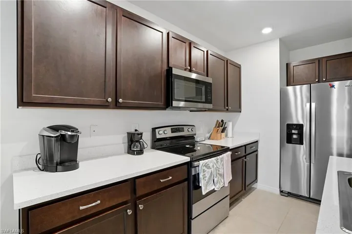 Kitchen featuring stainless steel appliances, dark brown cabinetry, light tile patterned flooring, light stone counters, and recessed lighting