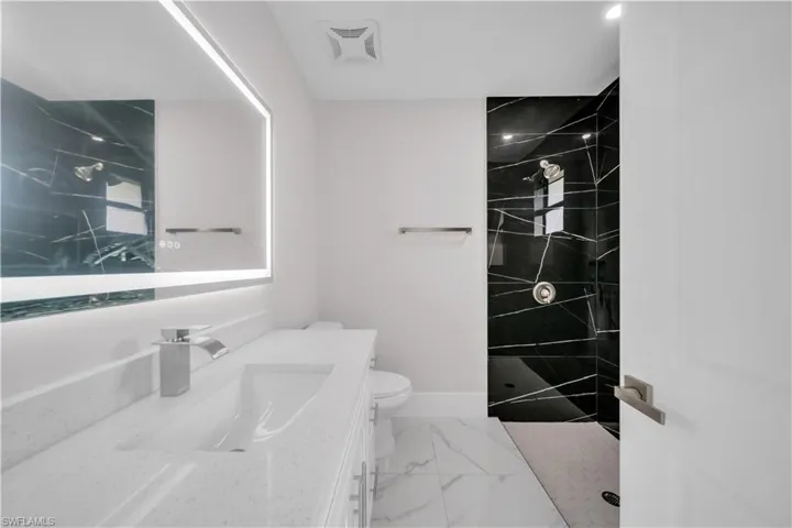 Walk-in shower with black tile surround, vanity with an integrated sink, illuminated mirror, and marble-look floor tile