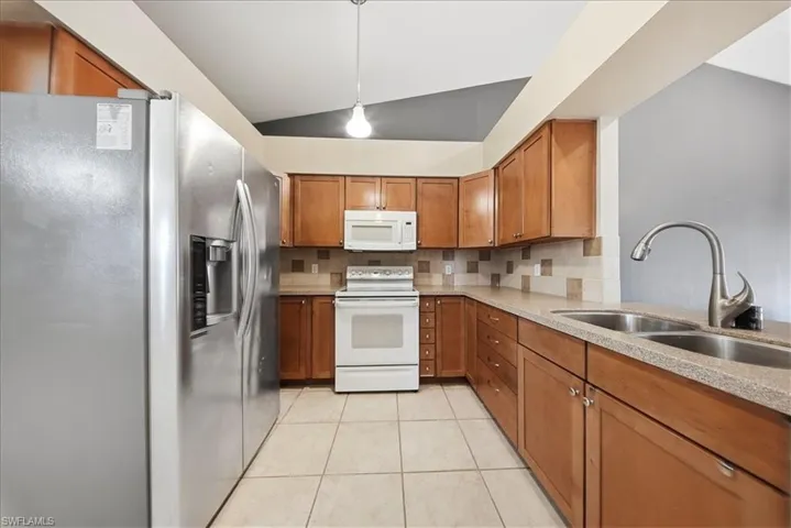 Kitchen featuring brown cabinets, white appliances, lofted ceiling, backsplash, and decorative light fixtures