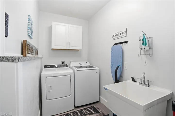 Dedicated laundry area featuring a utility sink with chrome faucet, granite countertop, built-in upper cabinetry, and wood-finish flooring - Virtually Edited Image