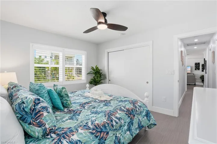 Bedroom featuring a ceiling fan with integrated lighting, a window with louvered shutters, light gray wall paint, and wood-finish flooring - Virtually Edited Image