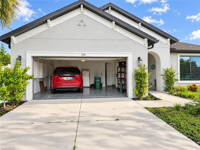 Stucco exterior with dark trim accents, attached garage with epoxy flooring, concrete driveway, arched entryway, and established landscaping