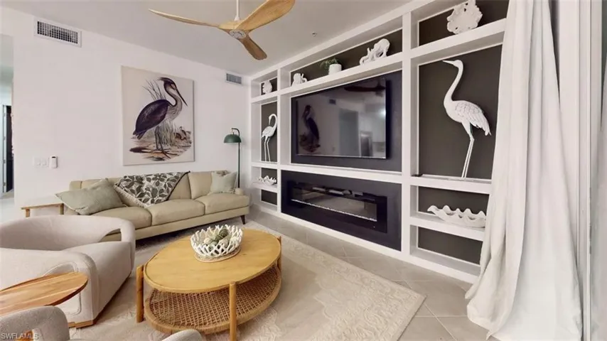 Living room with light tile patterned flooring, a ceiling fan, built in features, and a glass covered fireplace