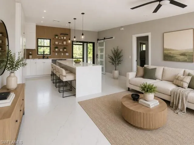 Open-concept living space with white tile flooring, a kitchen island with seating, white shaker cabinetry, a wood-paneled accent wall, and a ceiling fan