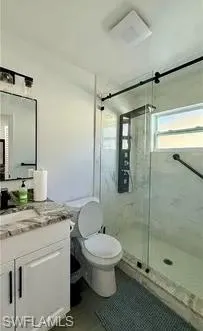 White vanity with a stone countertop, framed mirror, glass-enclosed shower, and a window