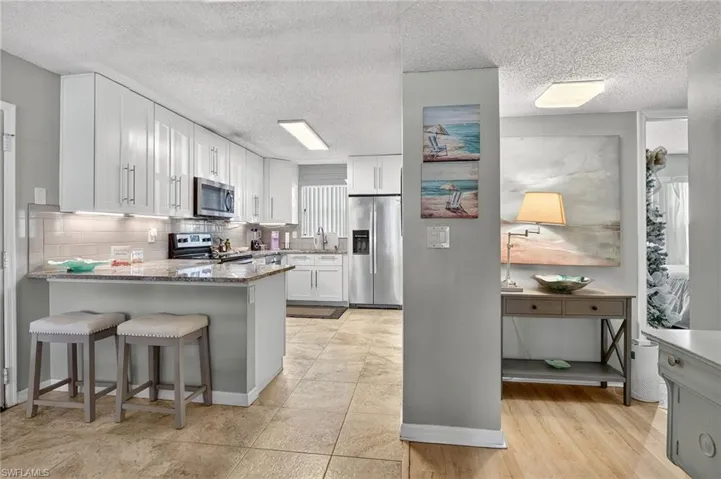 Kitchen with a textured ceiling, stainless steel appliances, light stone counters, white cabinetry, and a kitchen bar