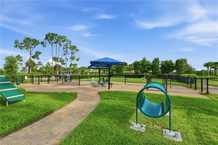 Dedicated outdoor amenity area featuring a paver pathway, green lawn, fenced enclosure, blue shade structure, and agility equipment