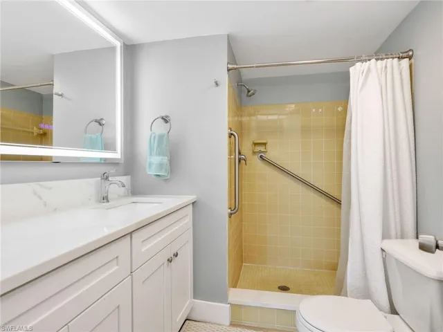 Bathroom featuring a vanity with a white countertop and integrated sink, a large illuminated mirror, a tile-surround shower with grab bars, and a toilet