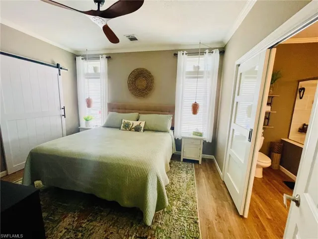 Bedroom with ceiling fan, a barn door, connected bathroom, light wood-type flooring, and crown molding