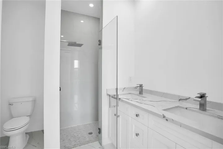 Double vanity featuring a white quartz countertop, undermount sinks with metallic faucets, a walk-in shower with large-format tile surround, rainfall showerhead, and a frameless glass enclosure