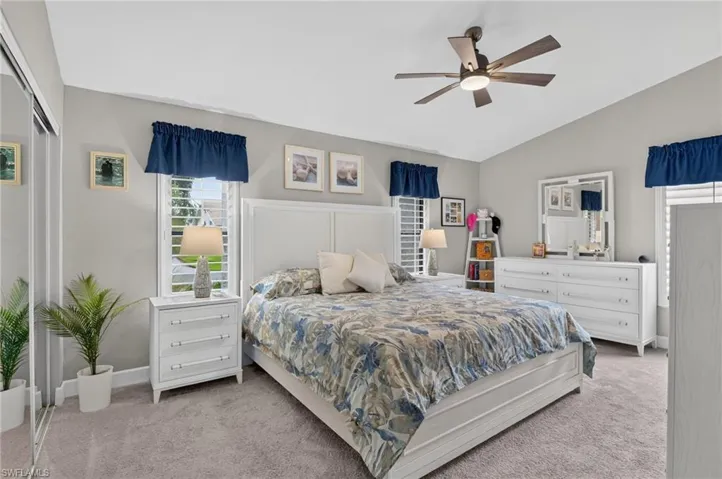 Vaulted ceiling with a ceiling fan, dual windows featuring plantation shutters, neutral-toned carpeting, and mirrored closet doors