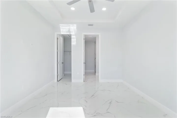 Unfurnished bedroom featuring a walk in closet, a tray ceiling, ceiling fan, recessed lighting, and light marble finish flooring