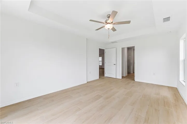 Unfurnished bedroom with a ceiling fan, visible vents, baseboards, light wood-type flooring, and a tray ceiling