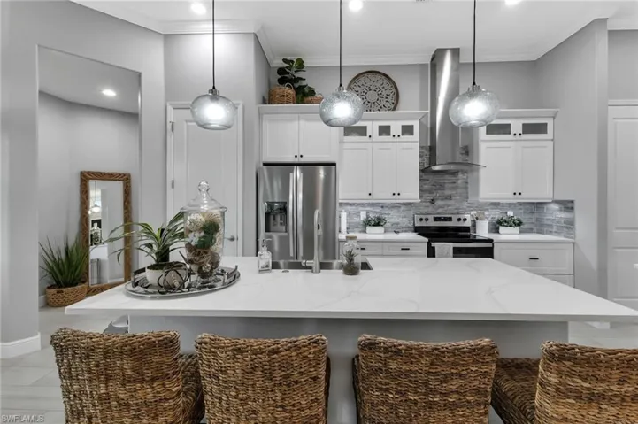 Kitchen featuring wall chimney range hood, a kitchen island with sink, backsplash, hanging light fixtures, and stainless steel appliances