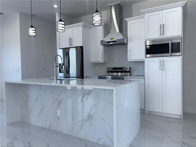 Kitchen with light stone countertops, white cabinetry, hanging light fixtures, wall chimney range hood, and appliances with stainless steel finishes