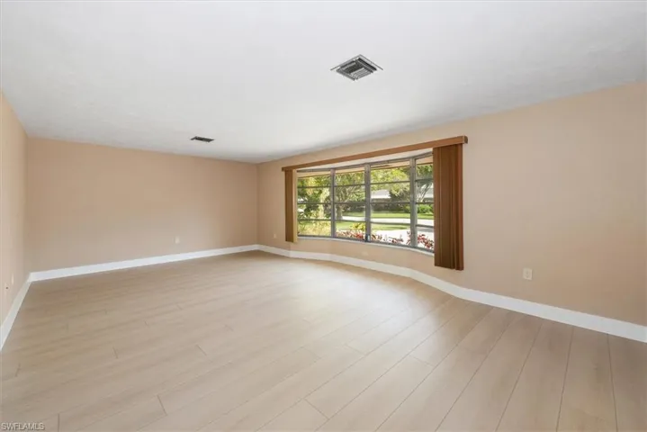 Spacious interior room featuring light wood-finish flooring, a neutral wall color, white baseboards, and a curved bay window with a wood valance and vertical blinds