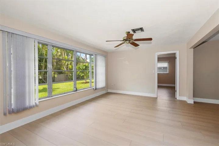 Spacious room featuring light wood-finish flooring, a large window overlooking a landscaped yard, and a ceiling fan with wood-tone blades