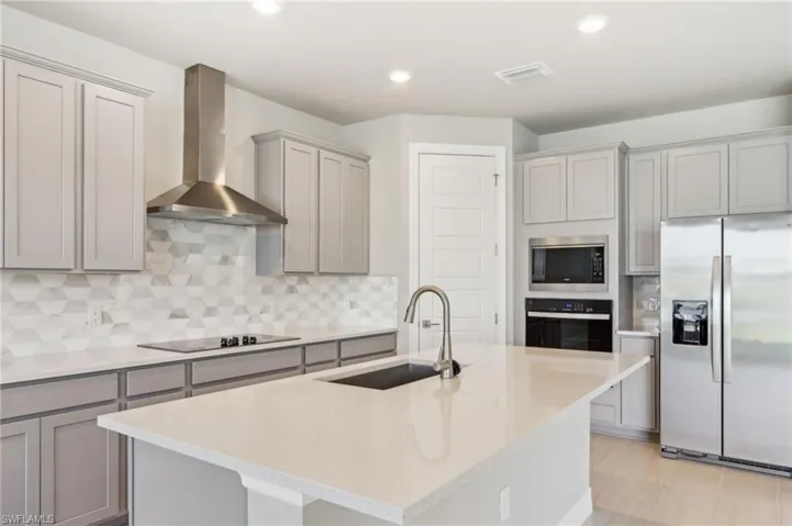 Kitchen with appliances with stainless steel finishes, a sink, wall chimney range hood, gray cabinetry, and a kitchen island with sink
