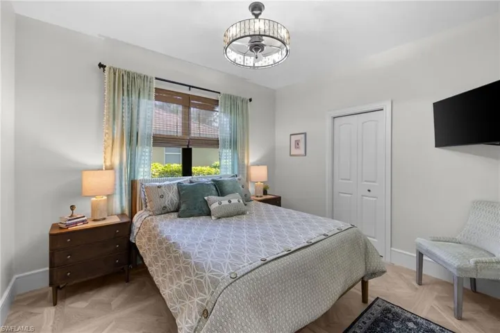 Bedroom featuring wood-finish herringbone flooring, a crystal-banded ceiling fan with integrated lighting, and a window with both woven wood shades and drapery