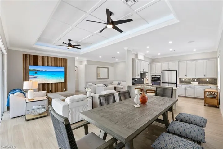 The open concept living area features tray ceilings with recessed lighting and ceiling fans, light wood-look flooring, and a kitchen with white cabinetry, stainless steel appliances, and a tiled backsplash